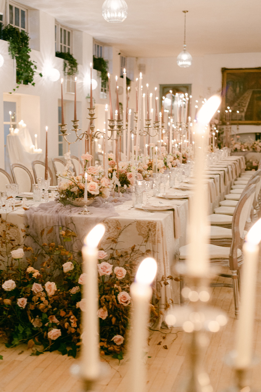 Elegant banquet table set with tall candles, floral arrangements, and white chairs in a softly lit room decorated with plants and paintings.