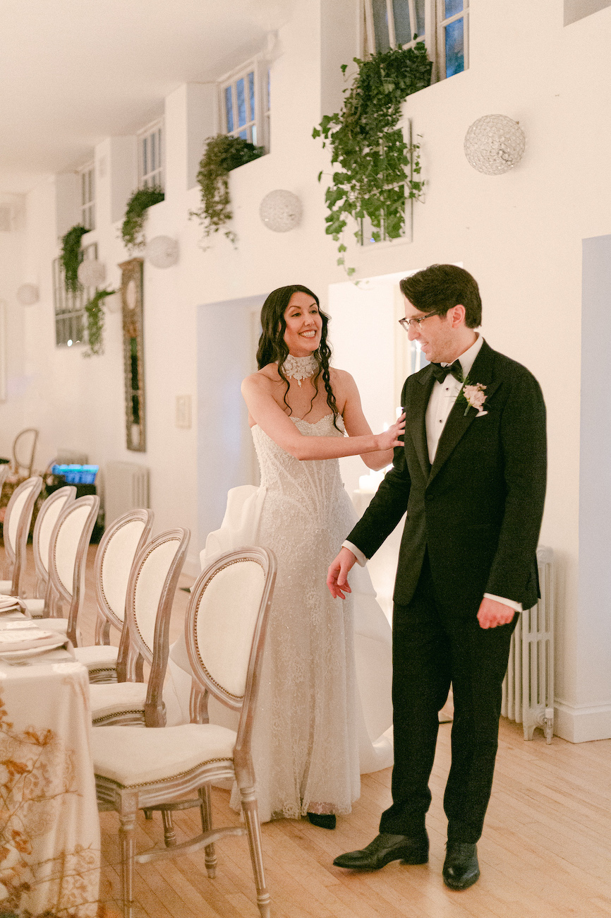 A woman in a white dress stands beside a man in a black tuxedo in a decorated room with elegant chairs and table settings.