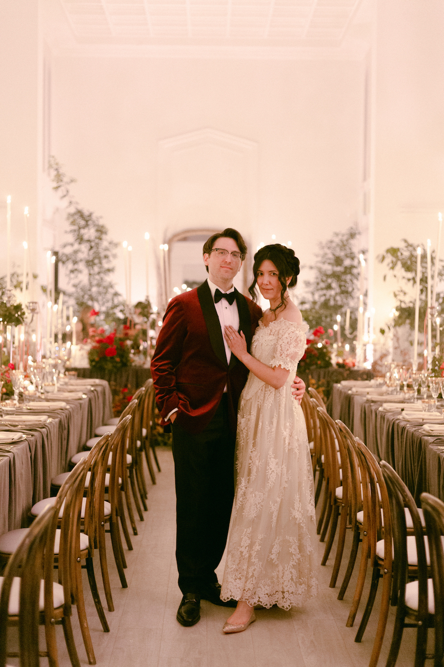 A couple dressed in formal attire stands together in the center of a decorated banquet hall lined with tables and chairs.
