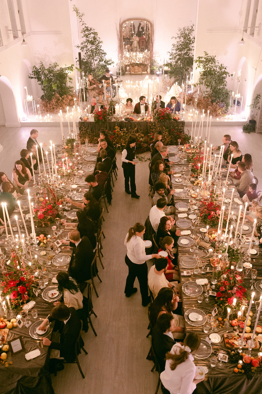 Guests seated at two long banquet tables decorated with many candles and flowers in a large, elegant hall.