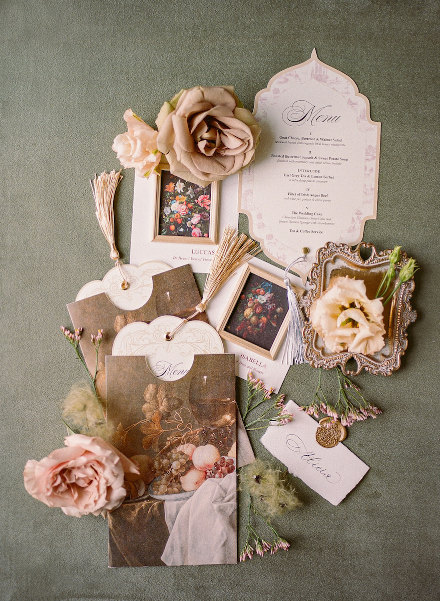 A flat lay of wedding stationery, including invitations, envelopes, floral photos, tassels, mirrors, tags, and artificial flowers arranged on a green surface.