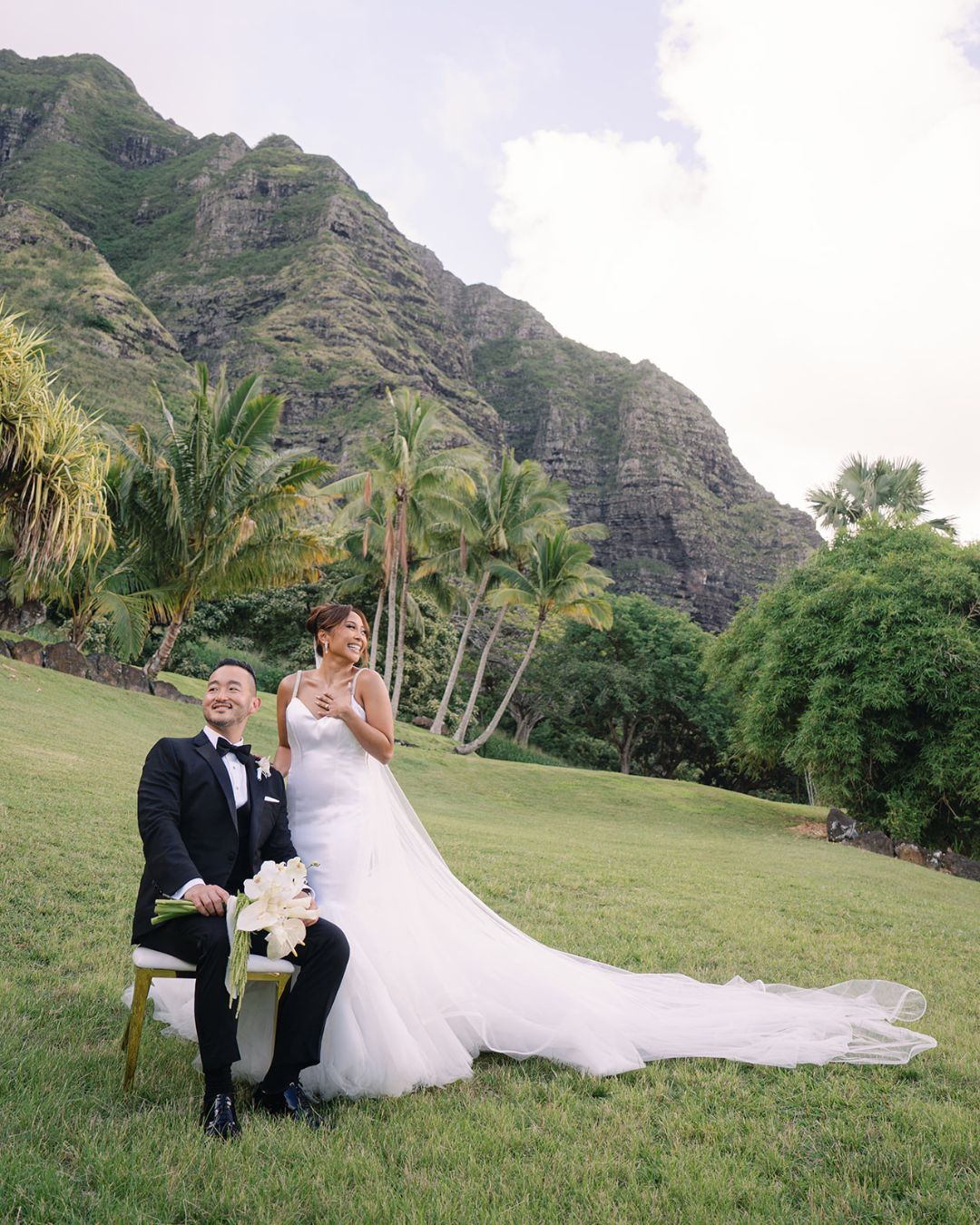 A bride in a white gown stands beside a seated groom in a black tuxedo on a grassy lawn with palm trees and mountains in the background.