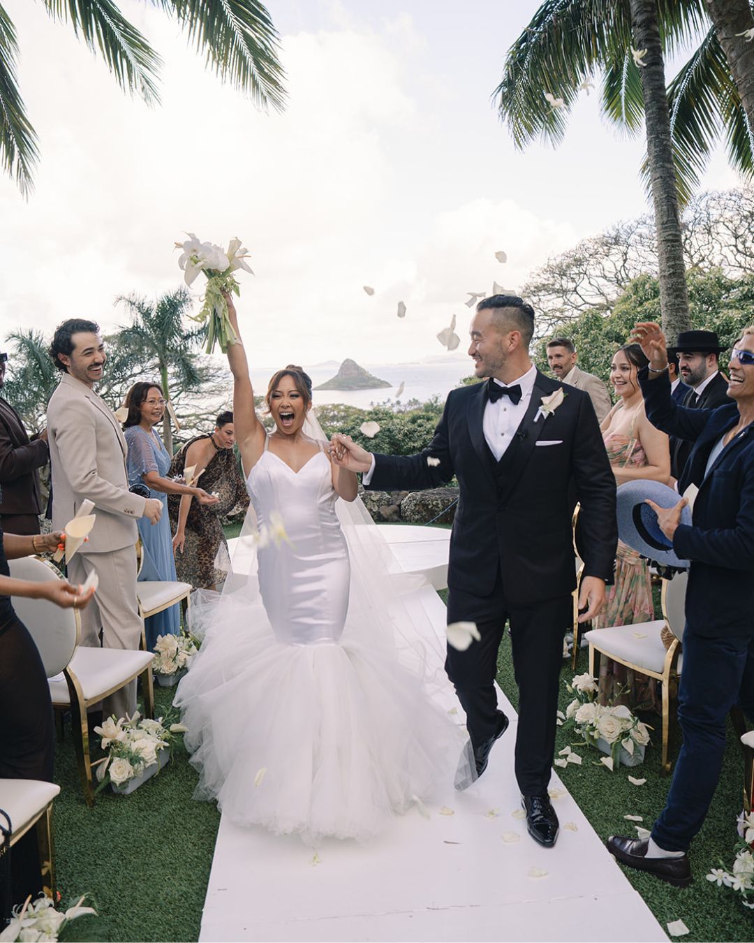 A bride and groom walk down an outdoor aisle, smiling and celebrating, as guests stand and clap. Palm trees and mountains are visible in the background.