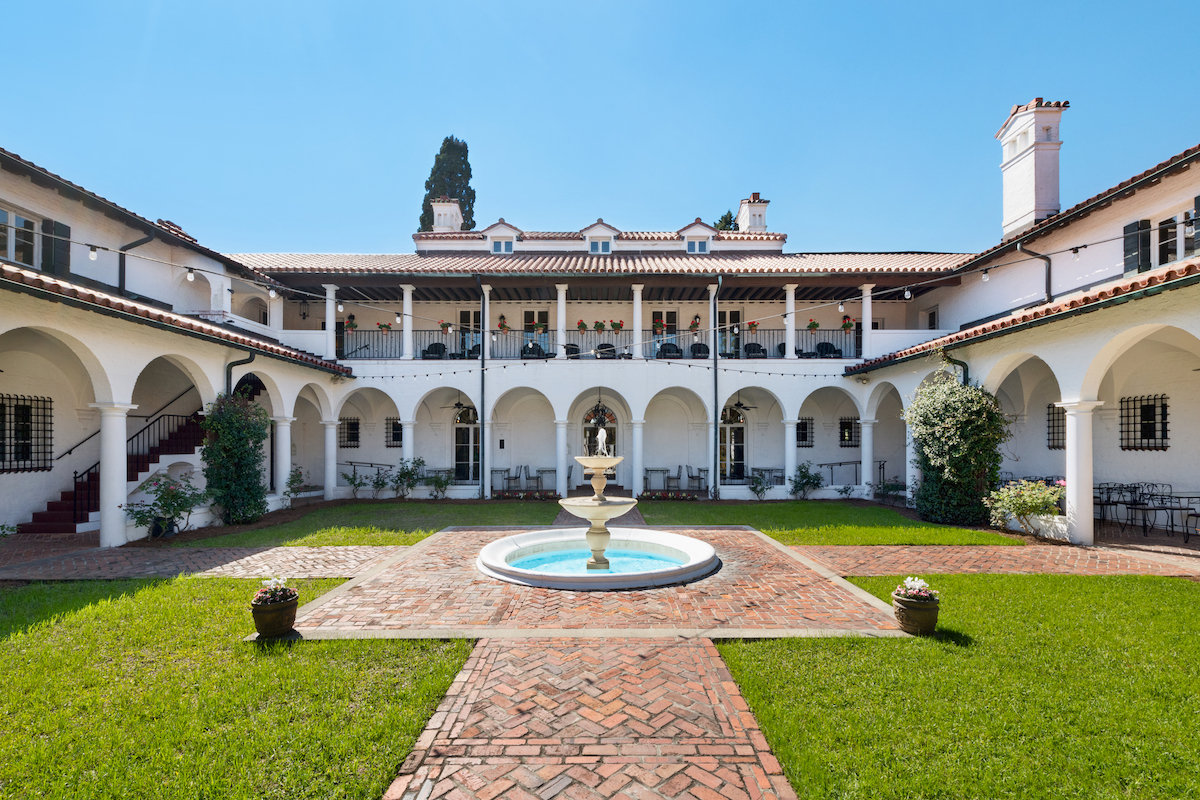 Sunny, enclosed courtyard with a brick walkway, central fountain, green lawn, white arches, and a two-story Mediterranean-style building in the background.