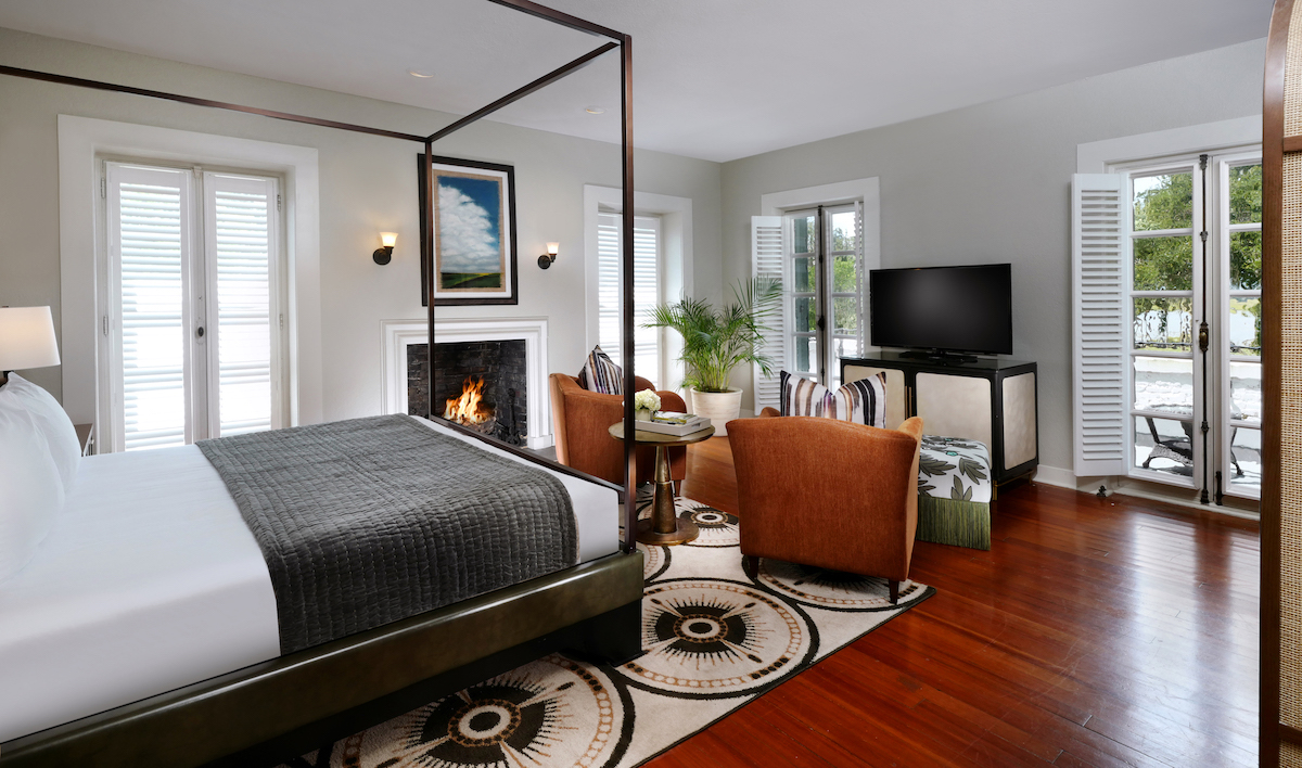 Modern bedroom with a four-poster bed, fireplace, TV, two armchairs, wood floor, and large windows with white shutters letting in natural light.