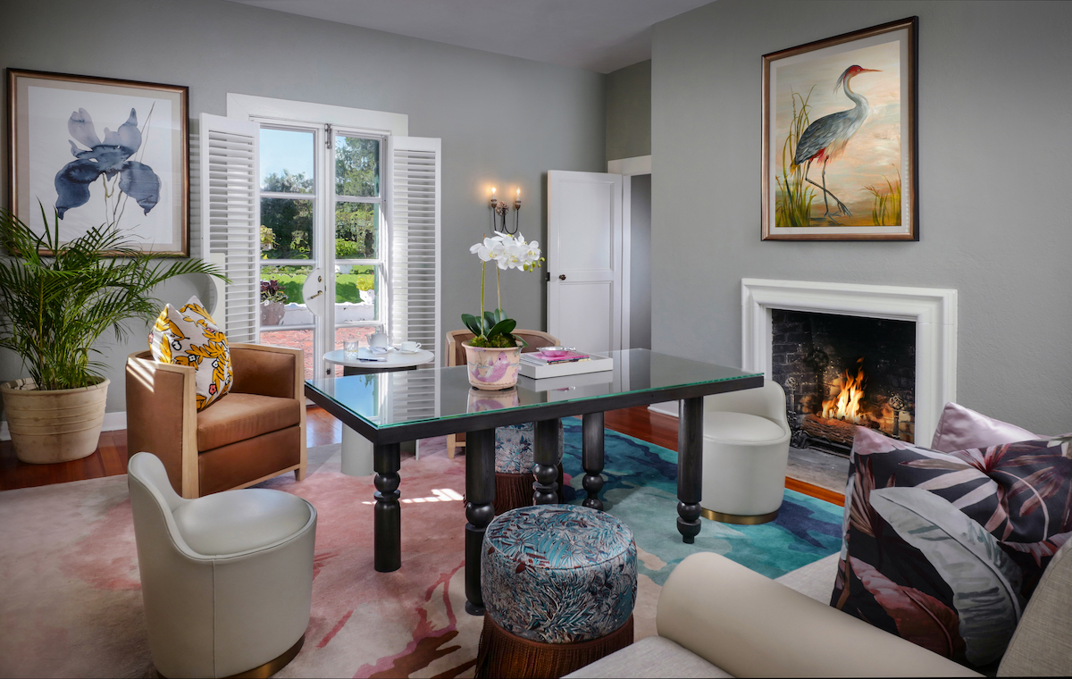 Modern living room with a glass table, fireplace, two brown chairs, white seating, plant, artwork, and open doors leading outside.