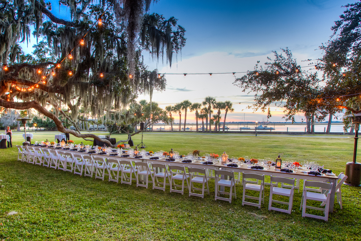 A long outdoor dining table with white chairs is set on grass under string lights, near trees, with a lake and sunset in the background.