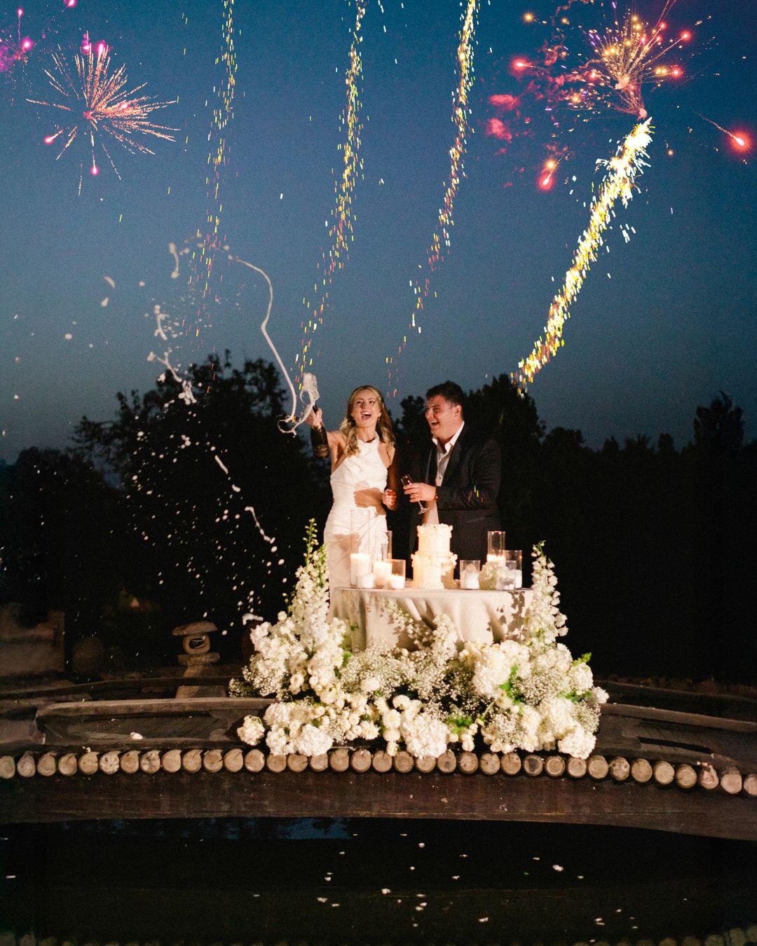 A bride and groom stand behind a wedding cake on a decorated table outdoors at night, with fireworks lighting up the sky above them.