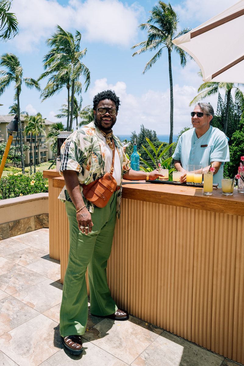 A man in green pants and a floral shirt stands at an outdoor bar, holding a drink, with a bartender nearby. Palm trees and blue sky are in the background.