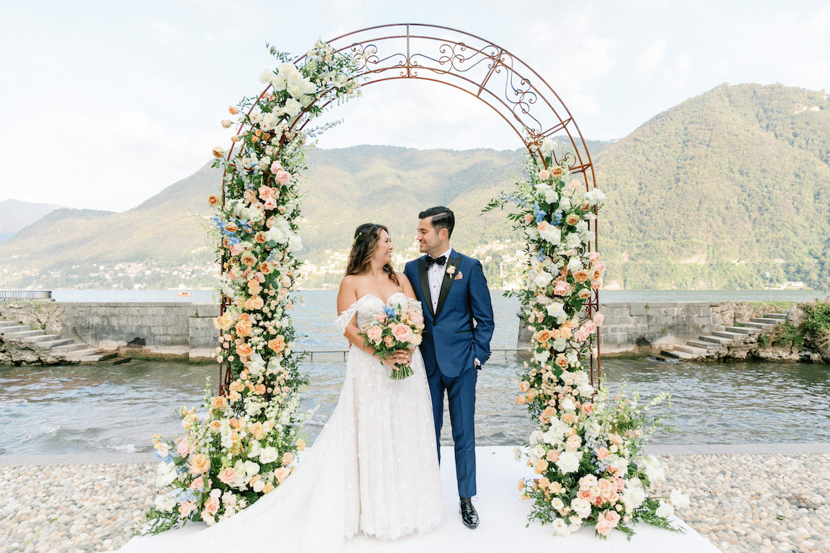 Bride and groom stand under a floral arch by a lakeside, facing each other, with mountains in the background.