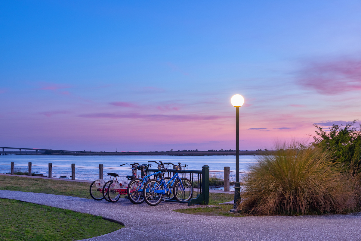 Several bicycles are parked near a pathway by the water at sunset, with a lit lamp post, grass, and distant bridge visible under a pink and blue sky.