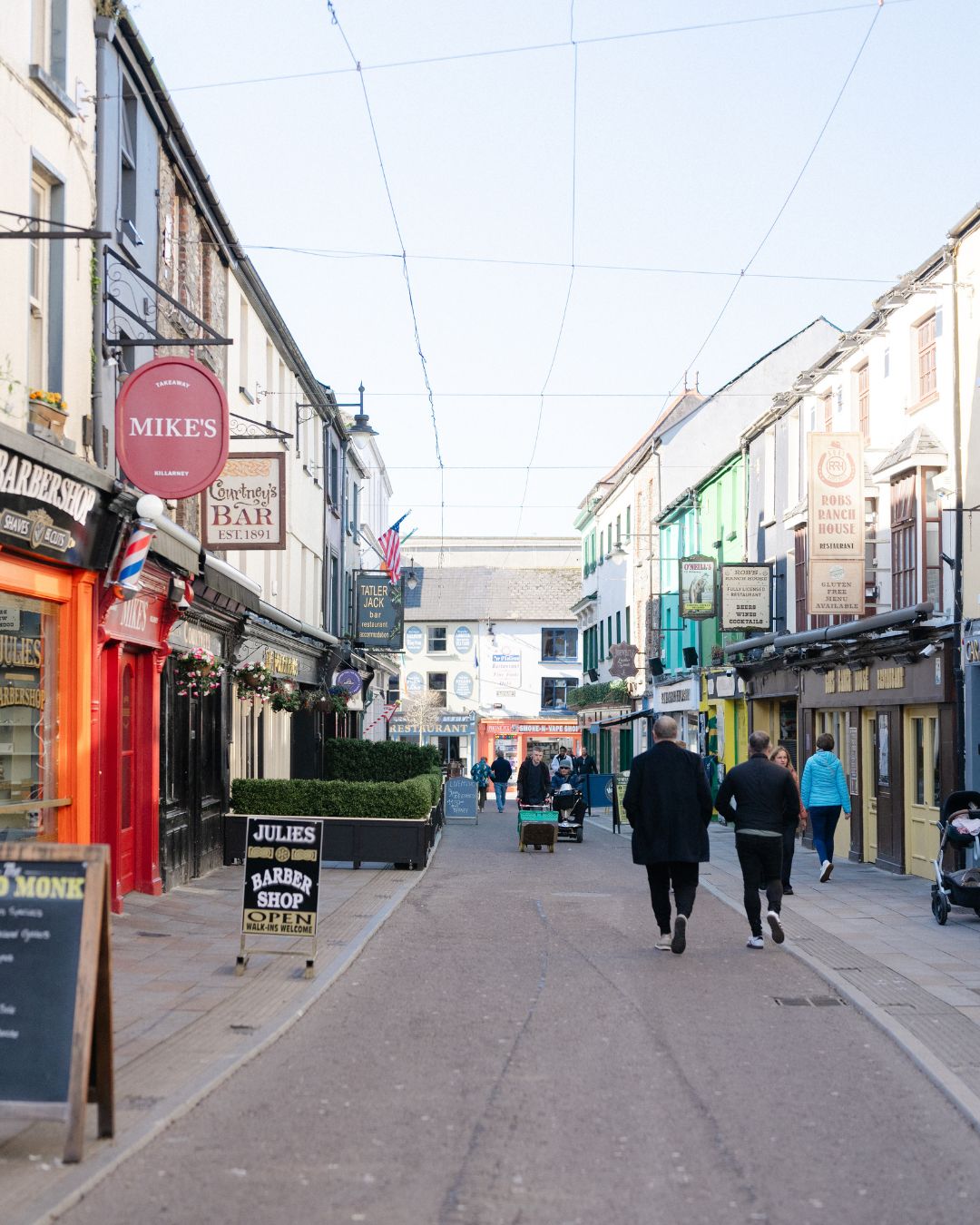 People walk down a narrow street lined with shops and colorful buildings on a clear day. Signs and sandwich boards are visible, and some pedestrians are in the distance.