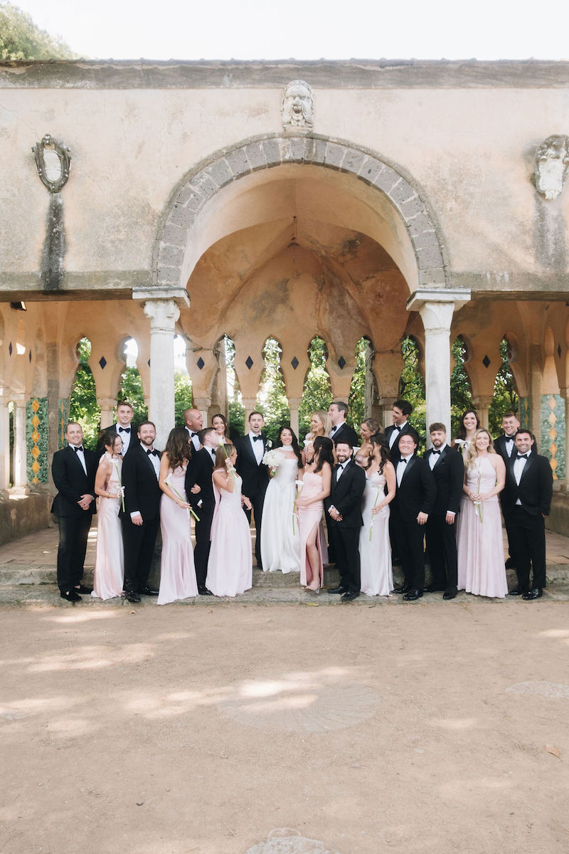 A wedding party poses outdoors in formal attire, with bridesmaids in pale dresses and groomsmen in black suits, standing before an arched stone structure.