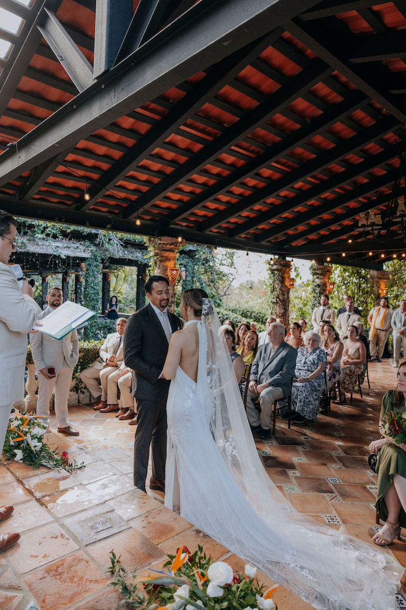 A bride and groom stand facing each other during a wedding ceremony under a covered outdoor patio, surrounded by seated guests.