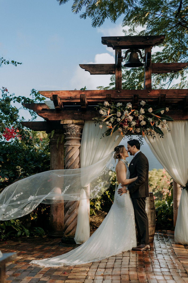A bride and groom stand under a wooden arch with flowers and draped fabric, embracing outdoors with the bride’s veil flowing in the wind.
