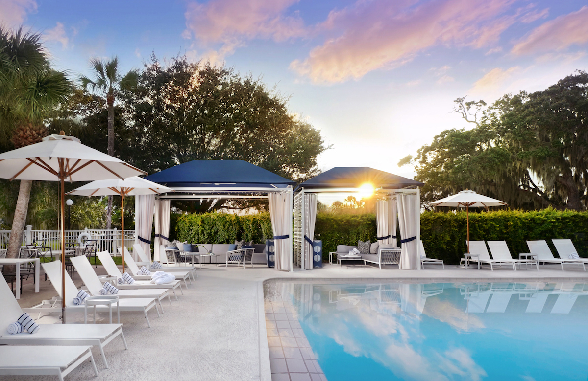 Outdoor pool area with lounge chairs, umbrellas, and cabanas, surrounded by trees and bushes at sunset under a partly cloudy sky.