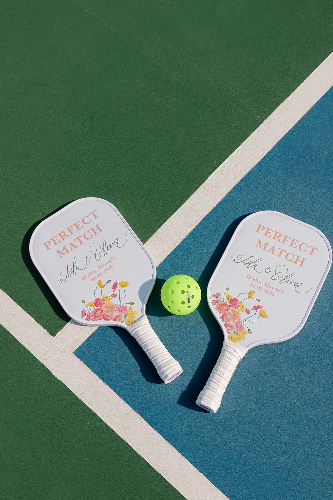 Two pickleball paddles with "Perfect Match" printed on them and a green ball are placed on a court near a white and blue line.