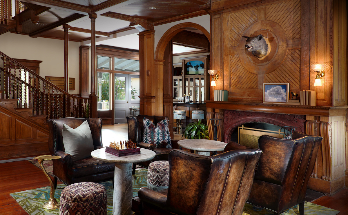 A wood-paneled living room features leather chairs, small round tables, a fireplace, a mounted animal head, and a staircase in the background.