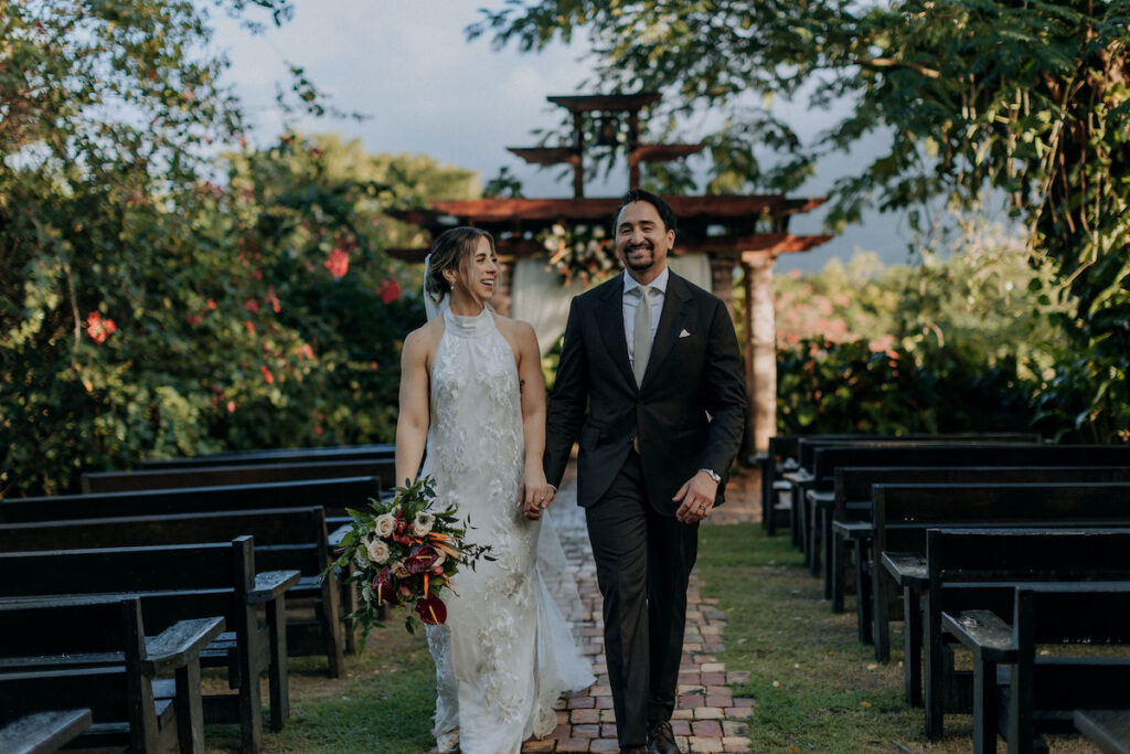 A bride in a white dress and a groom in a black suit walk hand in hand down an outdoor aisle, surrounded by greenery and empty benches.