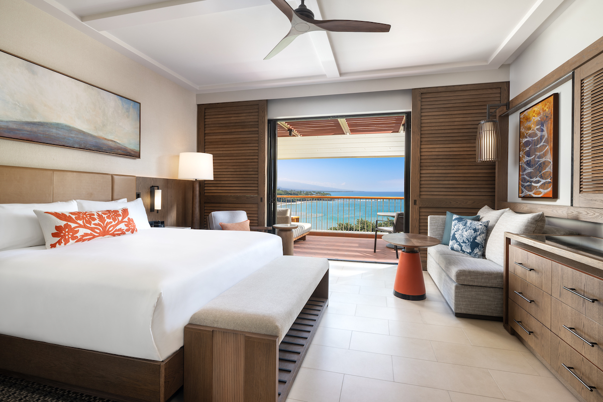 Modern hotel room with a large bed, sofa, and wooden decor, featuring open doors leading to a balcony with a view of the ocean.