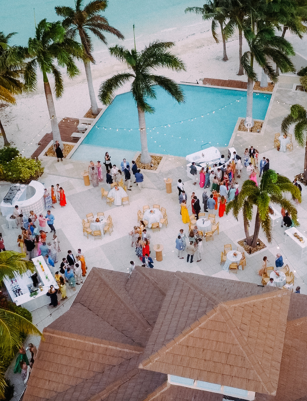 Aerial view of an outdoor event by a pool and beach, with guests gathered around tables, palm trees, and a coastal backdrop.