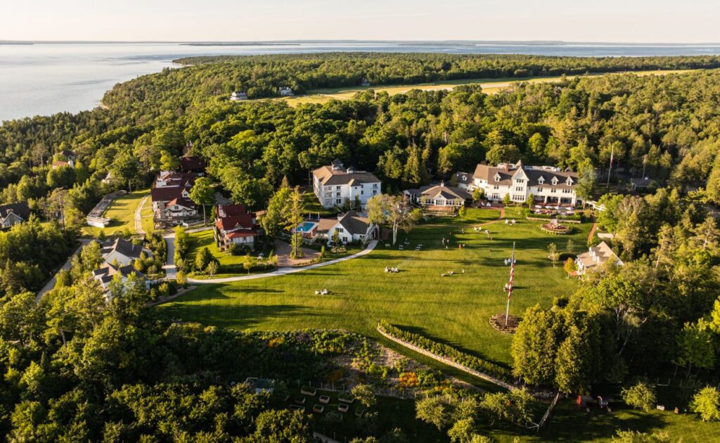 Aerial view of several large houses and open green lawns surrounded by dense forest near a body of water.