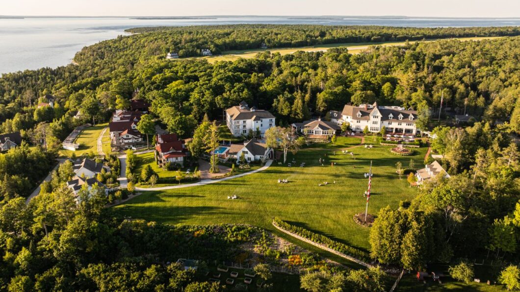 Aerial view of several large houses and open green lawns surrounded by dense forest near a body of water.