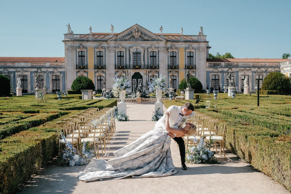 A couple poses in a dip at an outdoor wedding ceremony, surrounded by gold chairs and floral arrangements, in front of an ornate historic building.