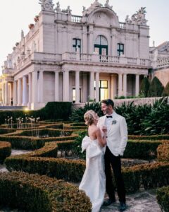 A bride and groom stand together in formal attire outside an ornate, classical building with manicured gardens and hedges in the foreground.
