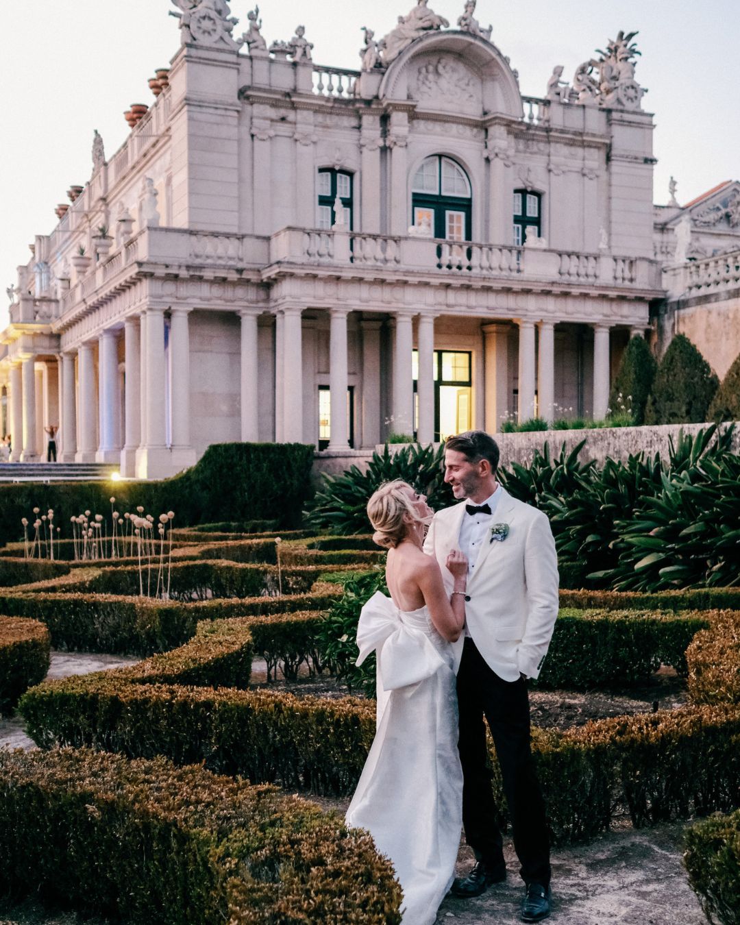 A bride and groom stand together in formal attire outside an ornate, classical building with manicured gardens and hedges in the foreground.