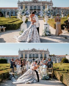 A bride and groom kiss at an outdoor wedding ceremony in front of a historic building, surrounded by guests, flowers, and manicured hedges.