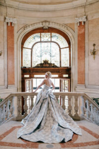 A person in a formal gown stands on an ornate staircase, facing a large arched window with stained glass in an elegant, historic building.