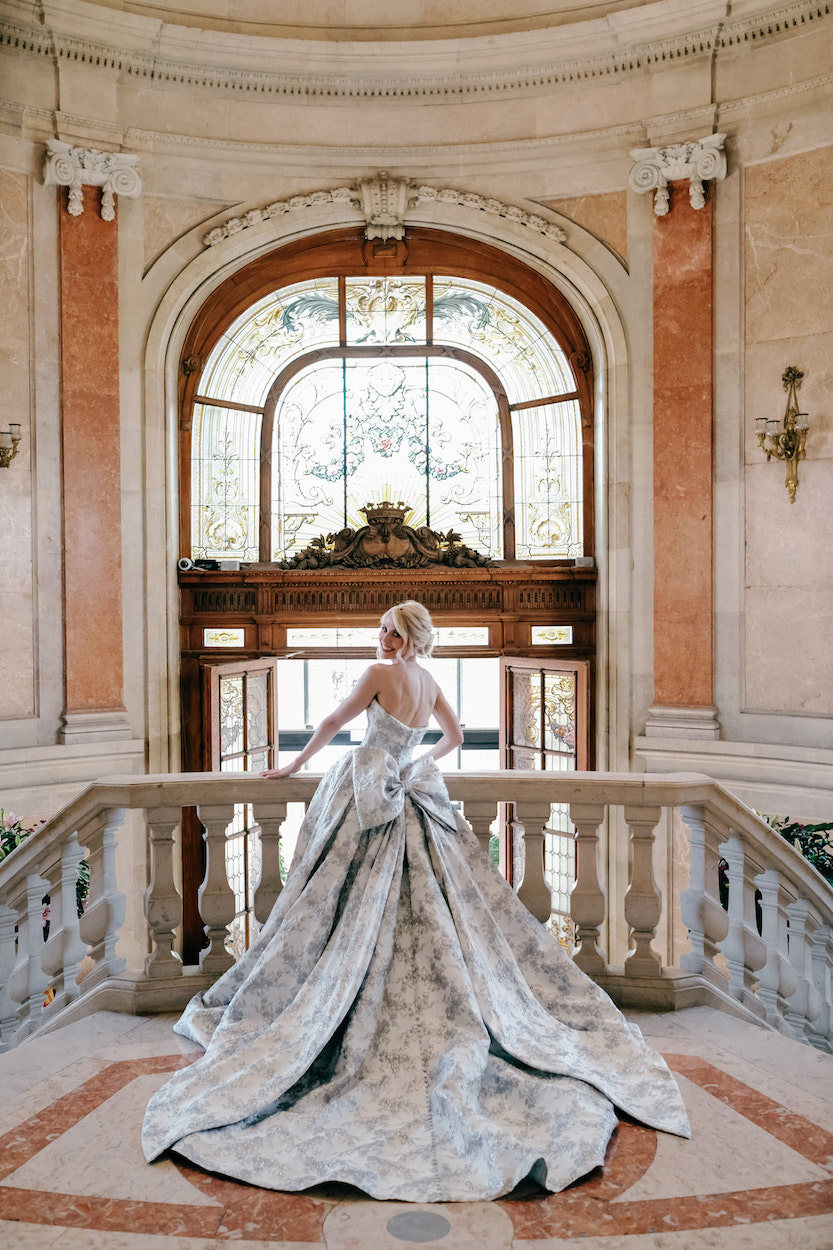 A person in a formal gown stands on an ornate staircase, facing a large arched window with stained glass in an elegant, historic building.