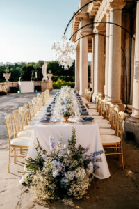 A long, elegant outdoor table set for a formal event with gold chairs, white tablecloth, blue accents, and floral arrangements, beside a stone building under a chandelier.