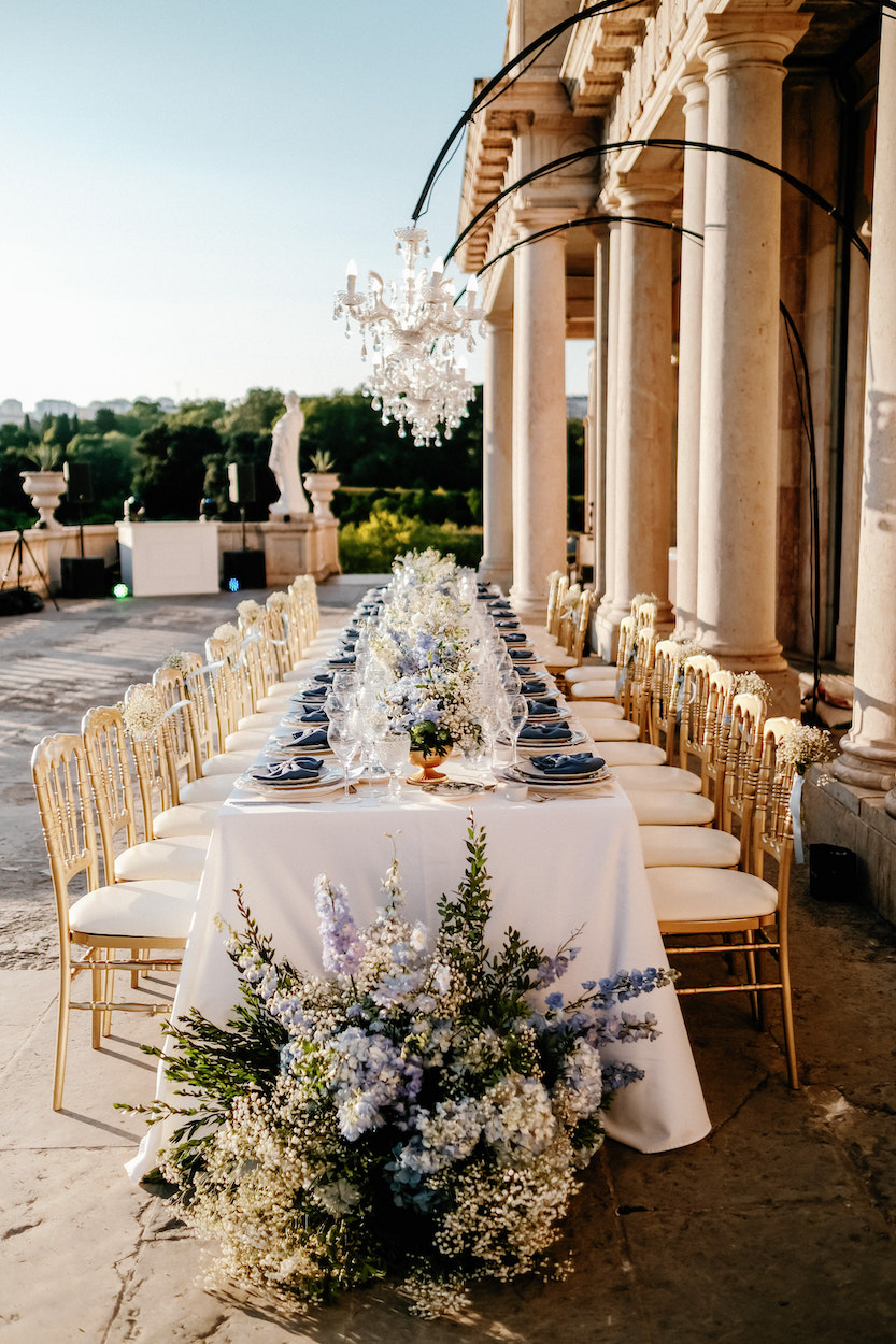 A long, elegant outdoor table set for a formal event with gold chairs, white tablecloth, blue accents, and floral arrangements, beside a stone building under a chandelier.