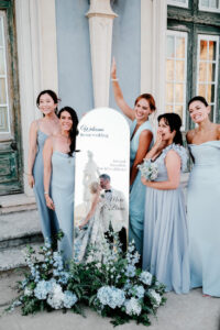 Five women in light blue dresses stand beside a decorative mirror sign with flowers at the base, posing and smiling outside a building with green-trimmed windows.