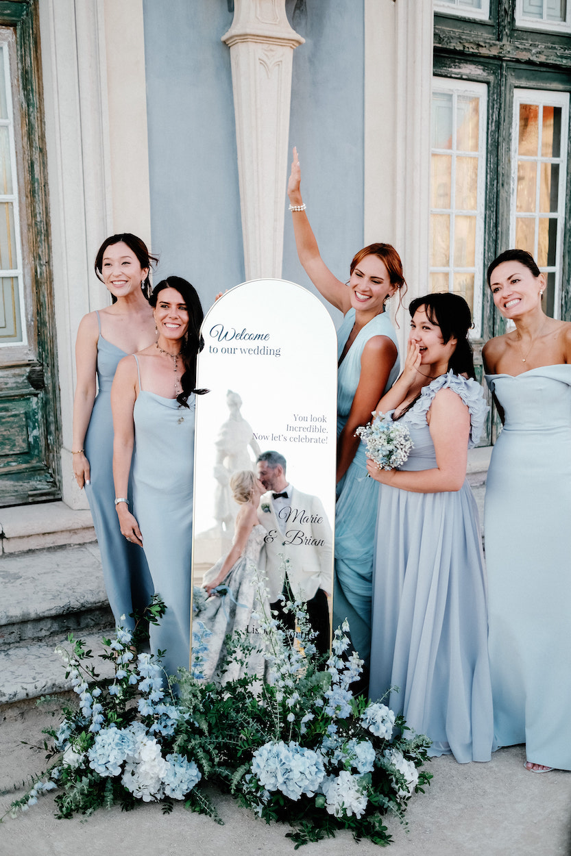 Five women in light blue dresses stand beside a decorative mirror sign with flowers at the base, posing and smiling outside a building with green-trimmed windows.