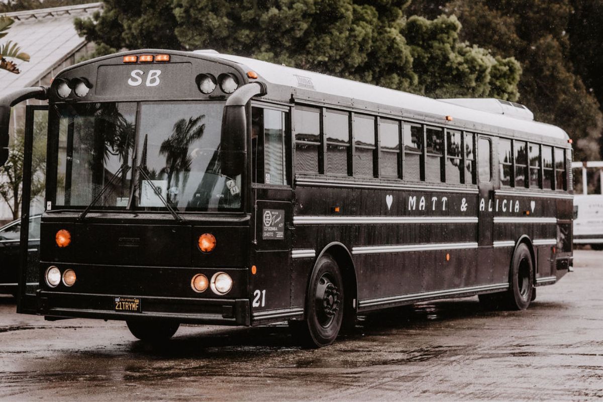 A black school bus with “SB” on the display is parked on a wet road, surrounded by trees and buildings in the background.