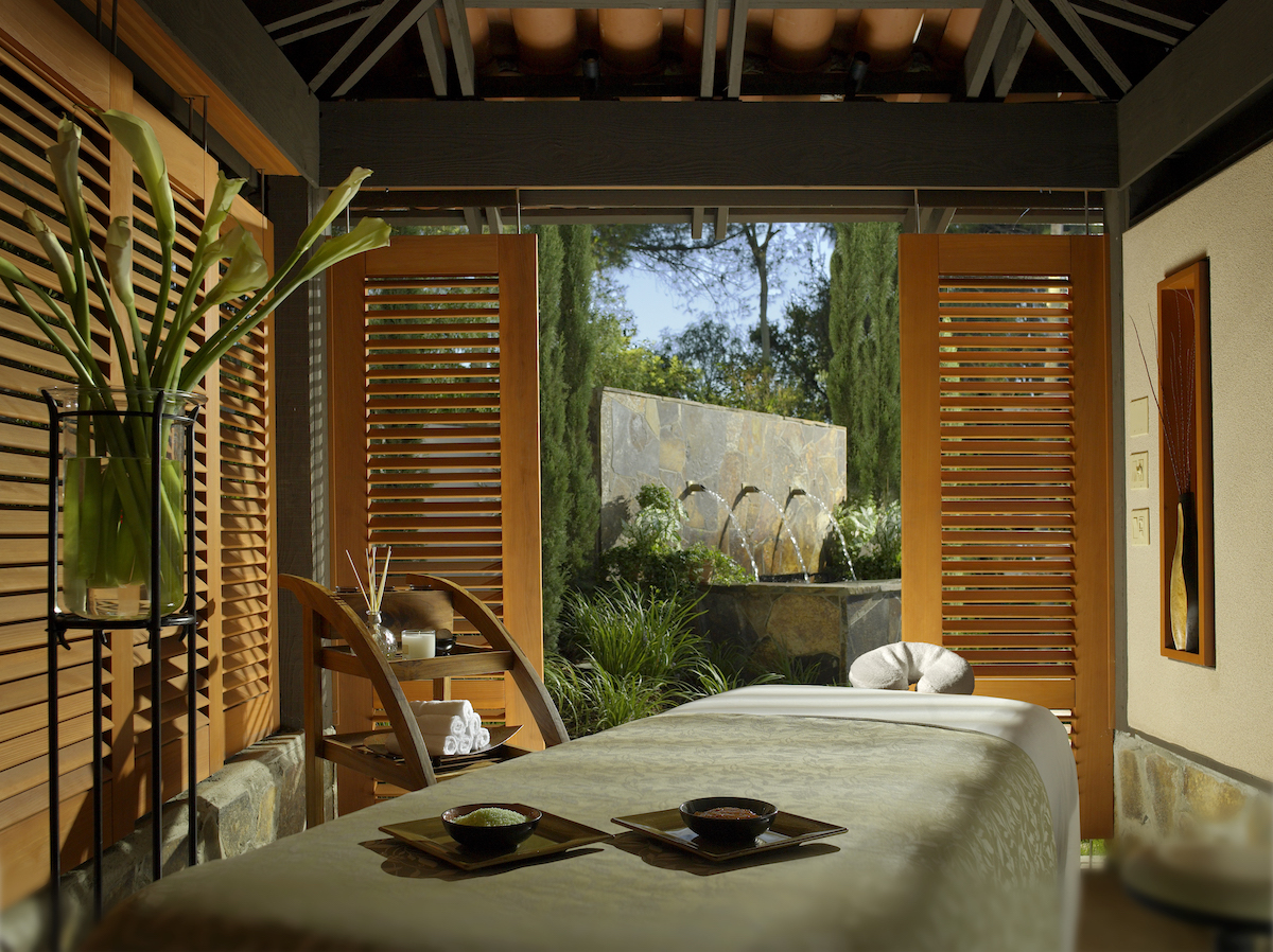 A massage table in a tranquil spa room with open wooden shutters, letting in natural light and a view of a garden and water fountain outside.