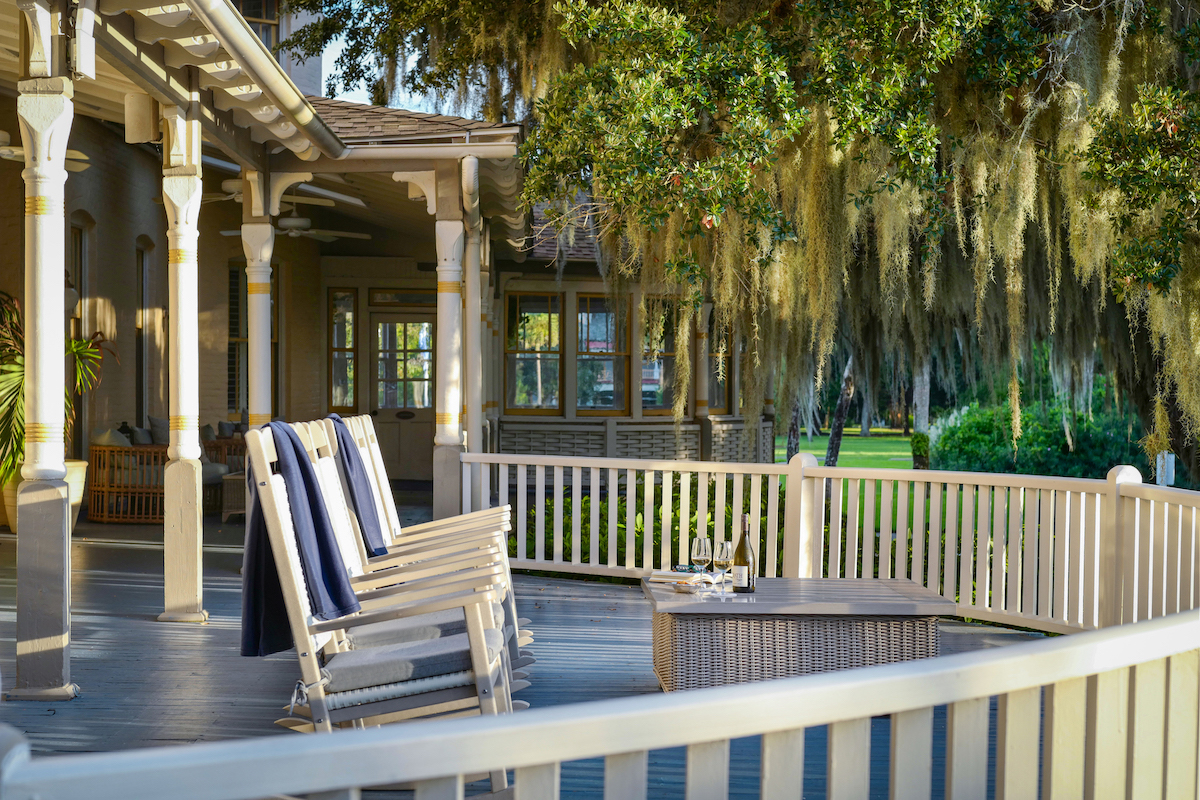 A wooden porch with a rocking bench, towels draped over the back, a wicker table, and trees with hanging moss in the background.