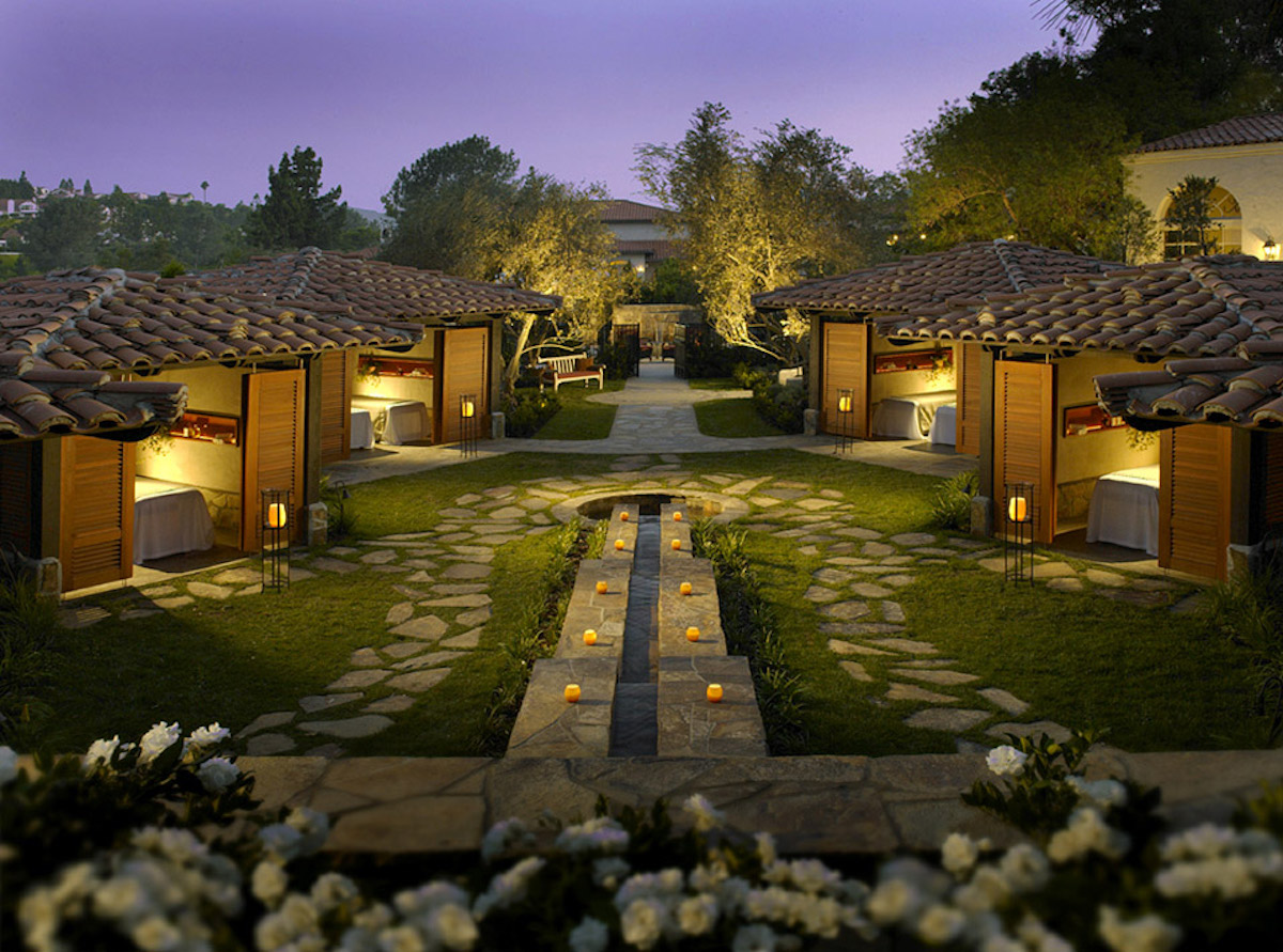 Outdoor view of a courtyard at dusk with stone paths, small cabanas, a central water feature, and lit lanterns, surrounded by trees and flowers.