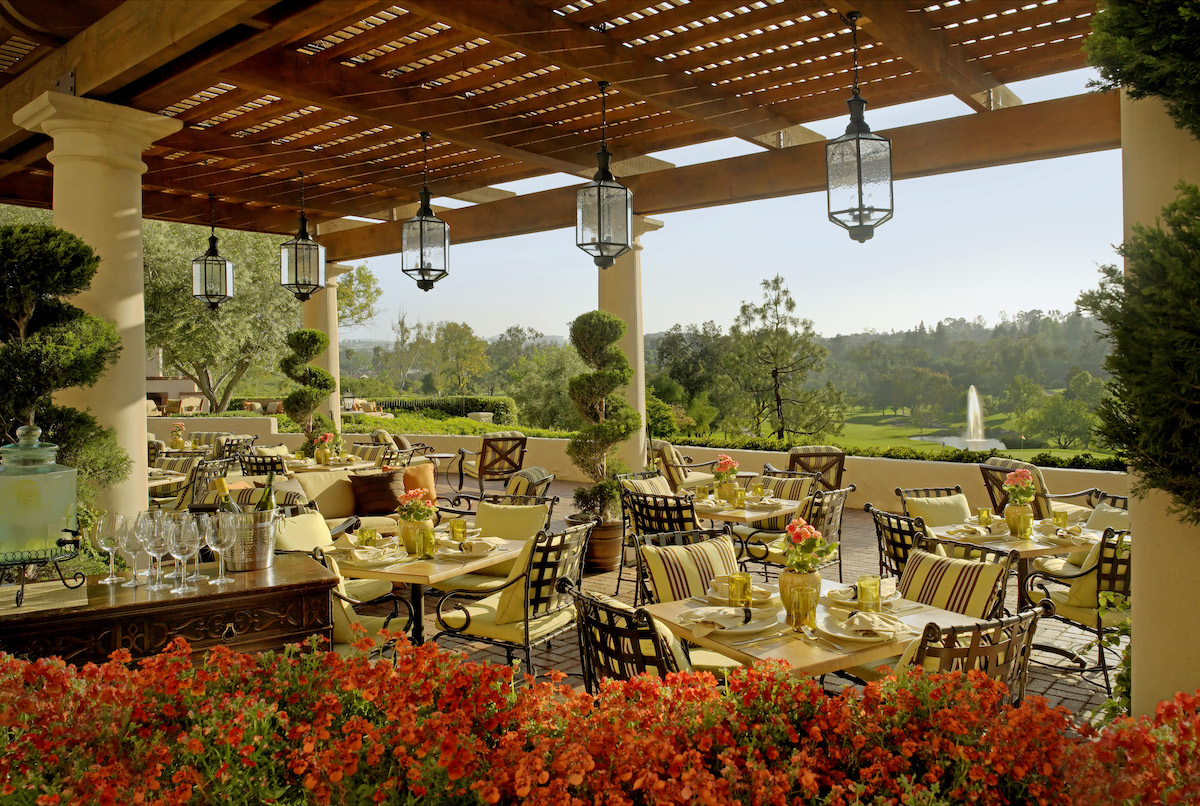 Outdoor restaurant patio with metal tables and chairs, set for dining under a wooden pergola, overlooking a landscaped garden with red flowers and a distant fountain.