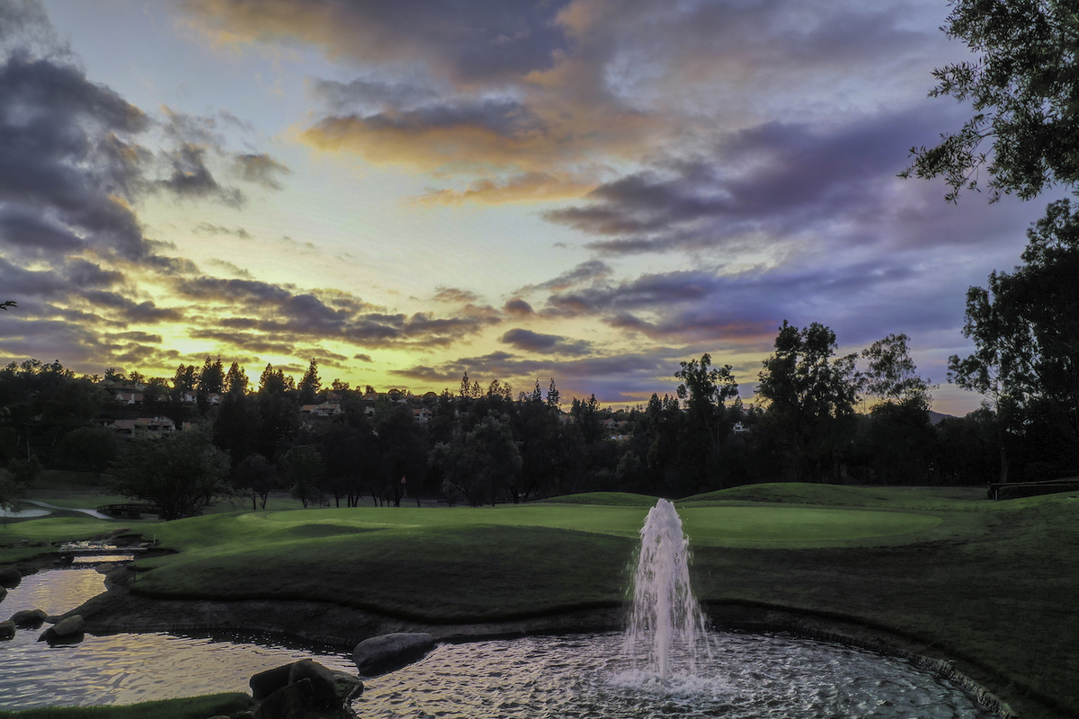 A golf course landscape at sunset with a small fountain in the foreground, surrounded by trees and a cloudy sky.
