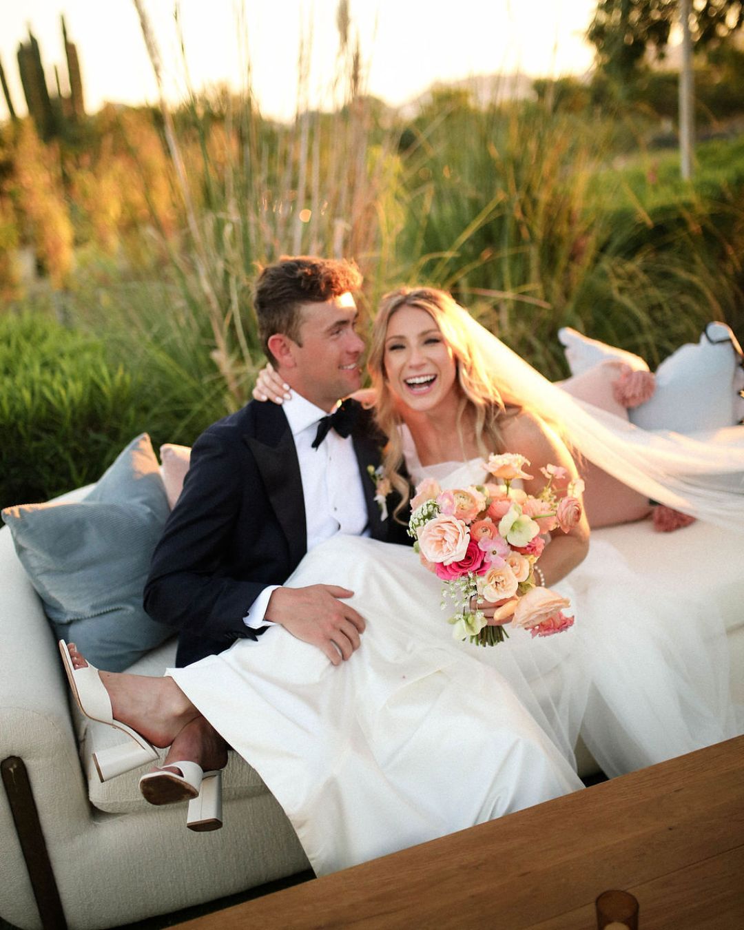 A bride and groom sit on an outdoor couch, smiling. The bride holds a bouquet of flowers and wears a white dress and veil; the groom is in a tuxedo. Sunlight illuminates the scene.