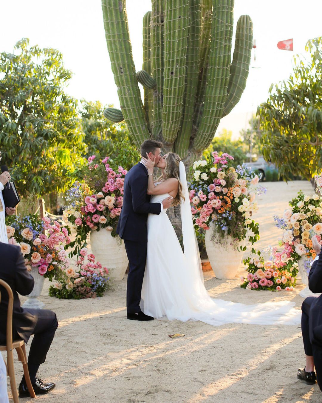 A bride and groom kiss at an outdoor wedding ceremony in front of a large cactus and colorful floral arrangements, with guests seated nearby.
