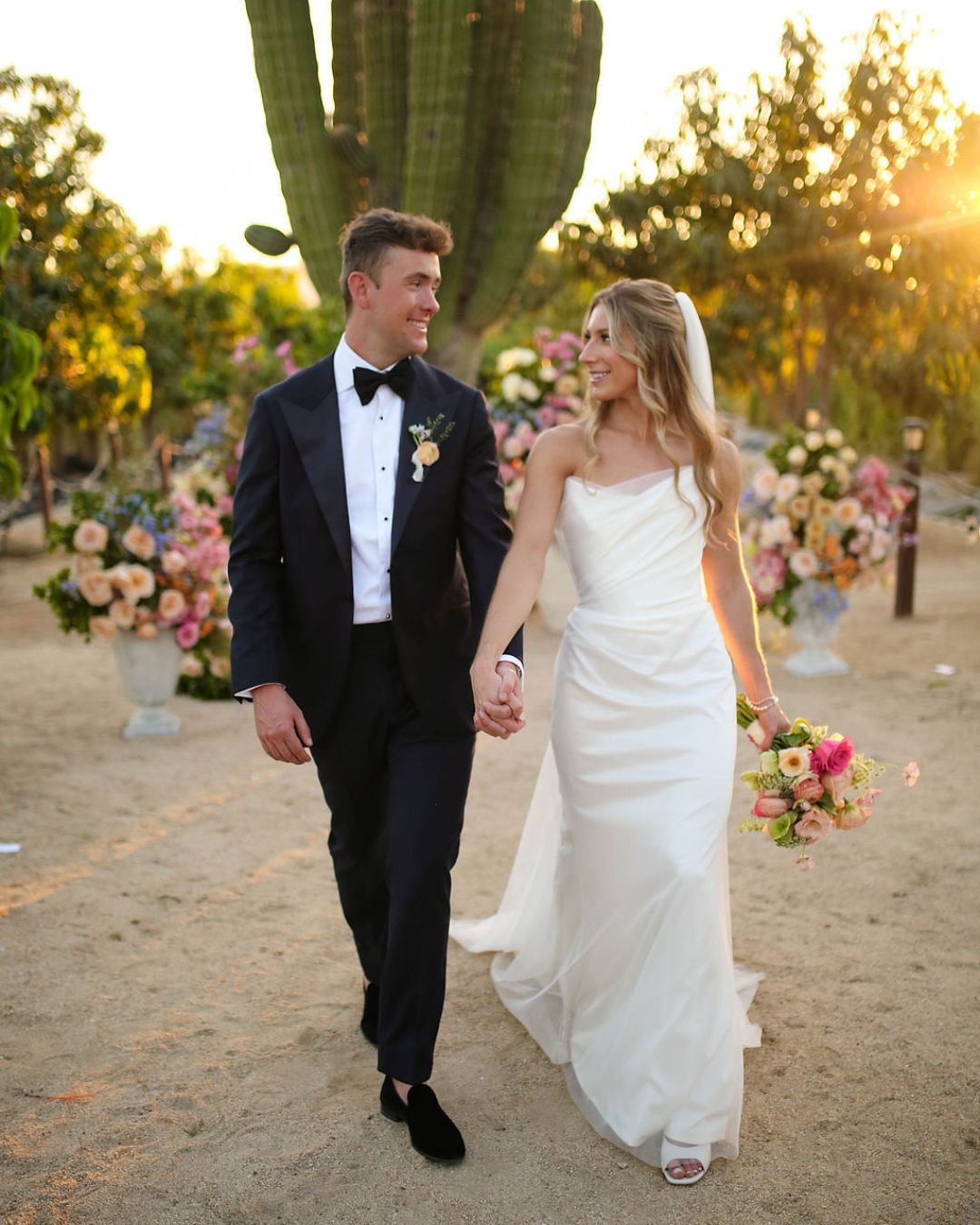 A bride and groom walk hand in hand outdoors at sunset, surrounded by flowers and cacti; both are smiling and dressed in formal wedding attire.