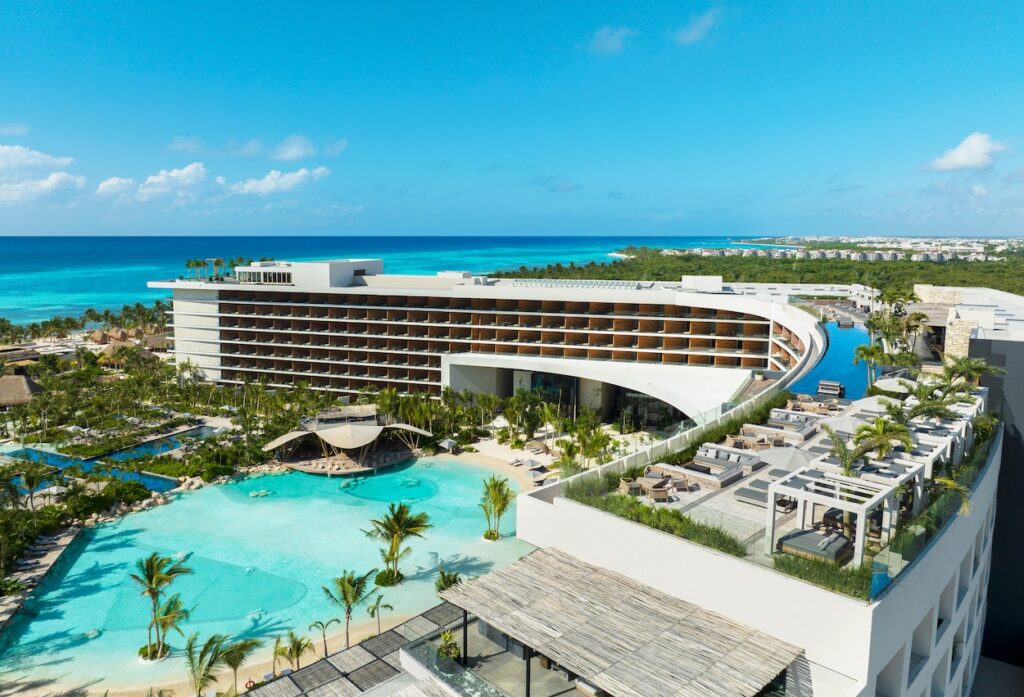 A modern beachfront resort with a curved multi-story building, outdoor pool, palm trees, and views of turquoise ocean under a clear blue sky.