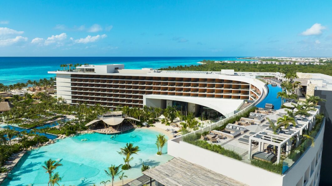A modern beachfront resort with a curved multi-story building, outdoor pool, palm trees, and views of turquoise ocean under a clear blue sky.