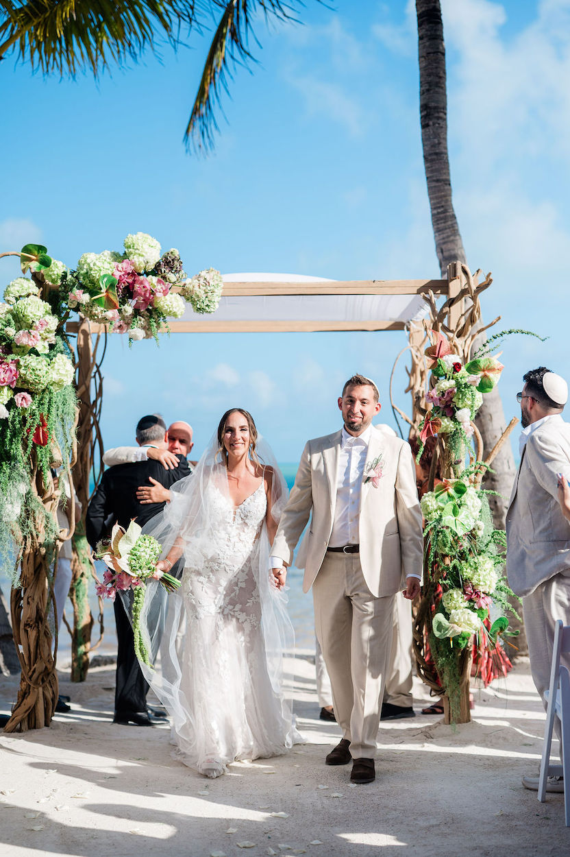 Bride and groom walk hand-in-hand under a decorated wedding arch on a sunny beach, with guests and floral arrangements visible around them.