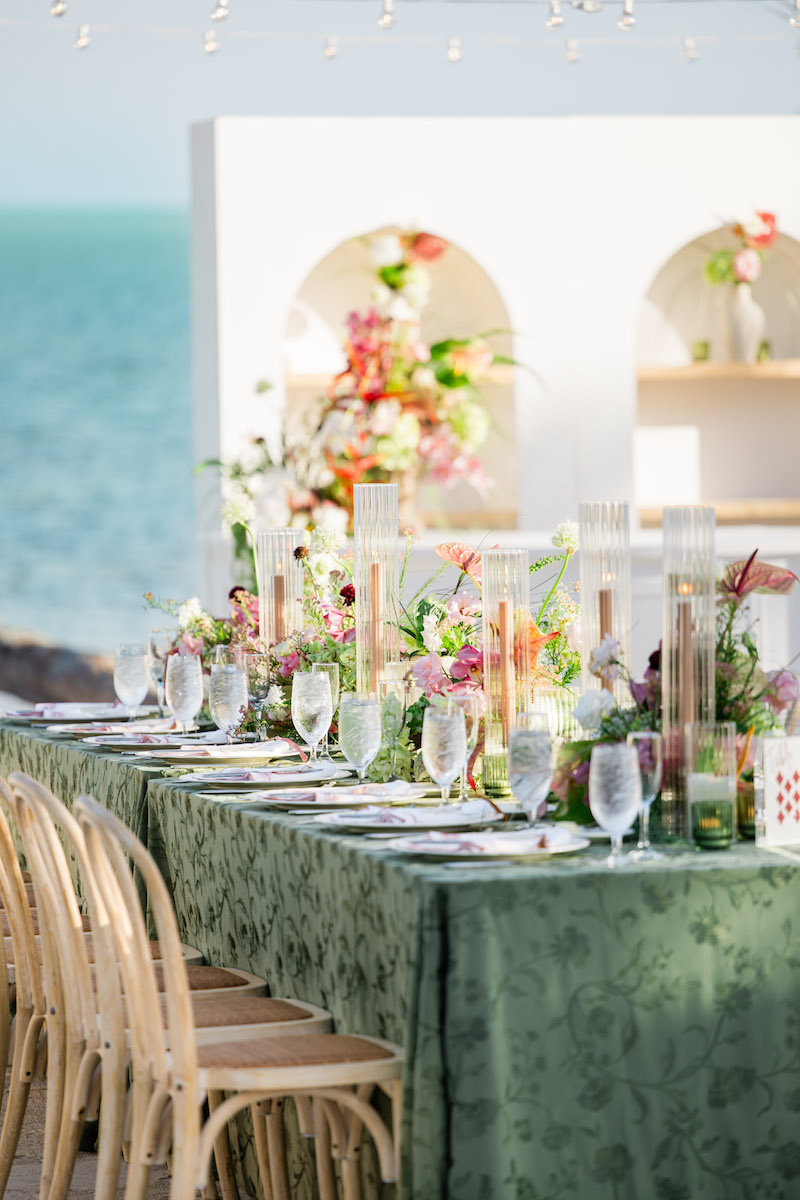 A long table with a green tablecloth is set for a formal event outdoors by the sea, decorated with flowers, glassware, and candles. Wooden chairs are arranged neatly along the sides.